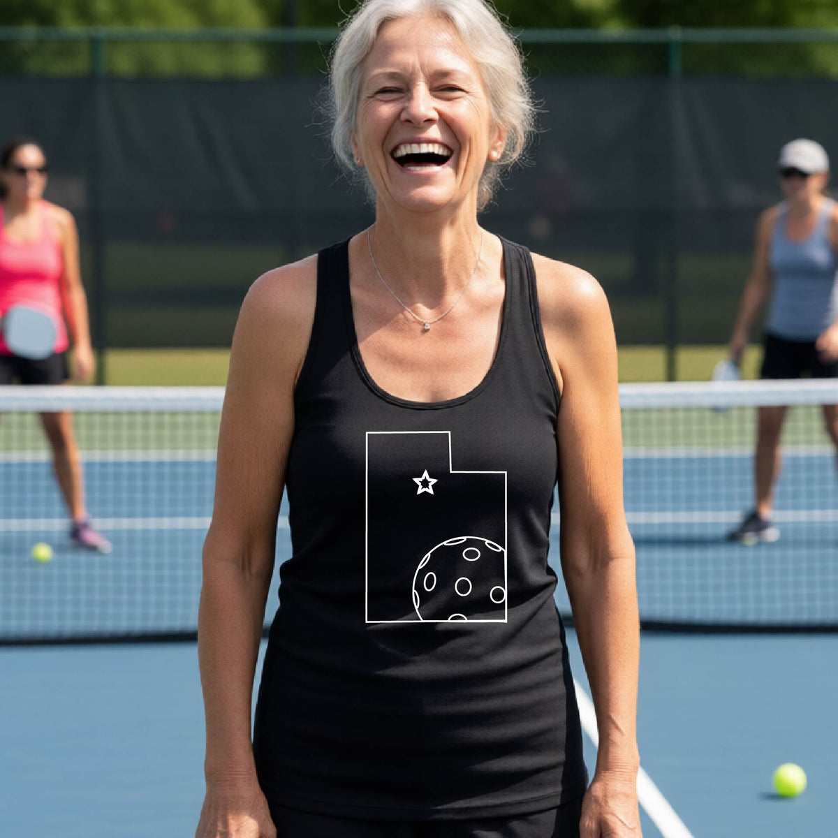 A close-up of a woman wearing a black racerback tank top, with a white graphic outline of the State of Utah , including an outline of a white pickleball in the lower right corner and a star outline in the center of the graphic, on the front of the hoodie.   

