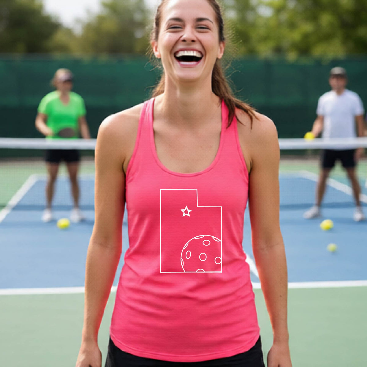A close-up of a woman wearing a coral pink racerback tank top, with a white graphic outline of the State of Utah , including an outline of a white pickleball in the lower right corner and a star outline in the center of the graphic, on the front of the hoodie.  
