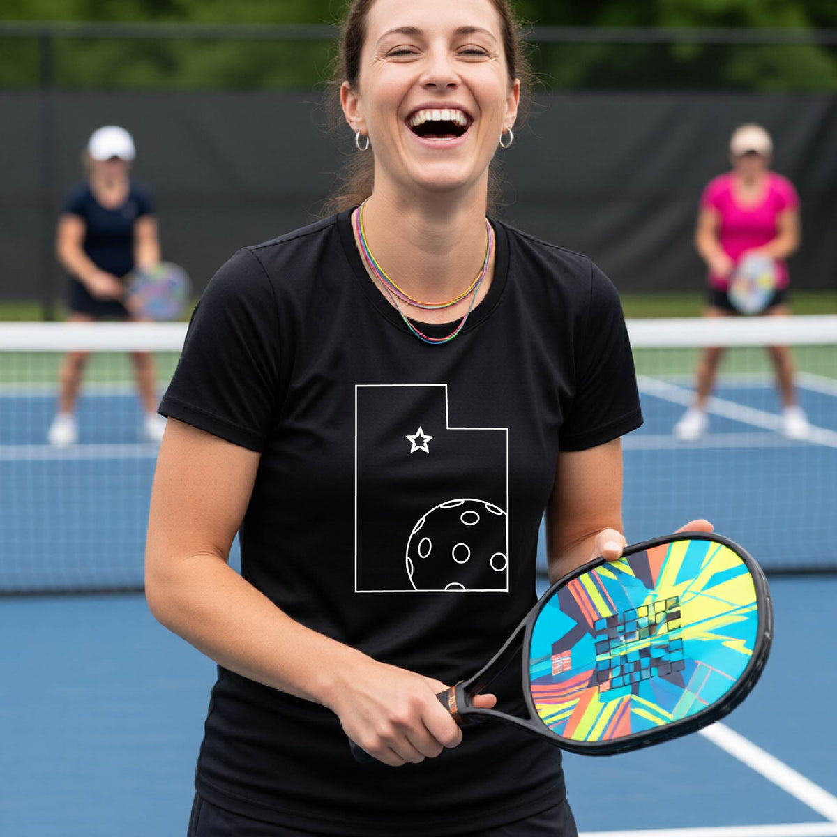 A close-up of a woman wearing a black short-sleeve, with a white graphic outline of the State of Utah , including an outline of a white pickleball in the lower right corner and a star outline in the center of the graphic, on the back of the shirt.   
