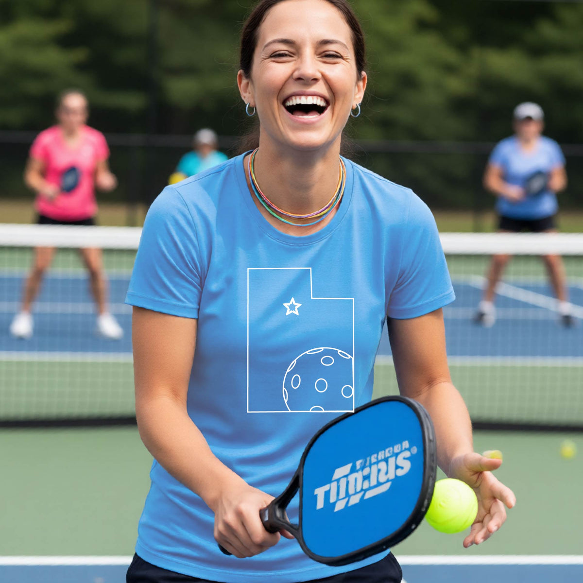 A close-up of a woman wearing a light blue short-sleeve, with a white graphic outline of the State of Utah , including an outline of a white pickleball in the lower right corner and a star outline in the center of the graphic, on the back of the shirt.   