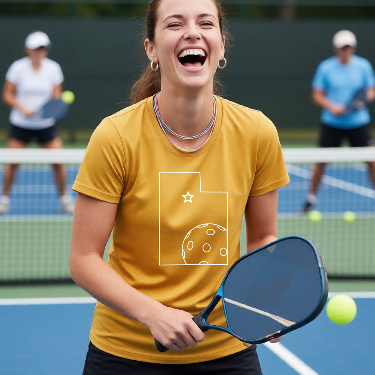 A close-up of a woman wearing a mustard yellow gold short-sleeve, with a white graphic outline of the State of Utah , including an outline of a white pickleball in the lower right corner and a star outline in the center of the graphic, on the back of the shirt.   