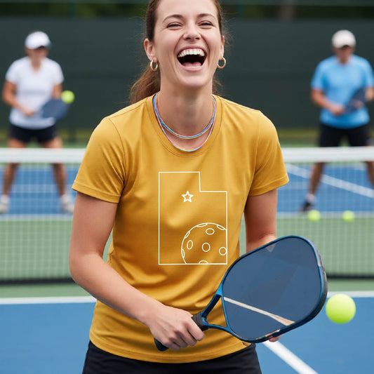 A close-up of a woman wearing a mustard yellow gold short-sleeve, with a white graphic outline of the State of Utah , including an outline of a white pickleball in the lower right corner and a star outline in the center of the graphic, on the back of the shirt.   