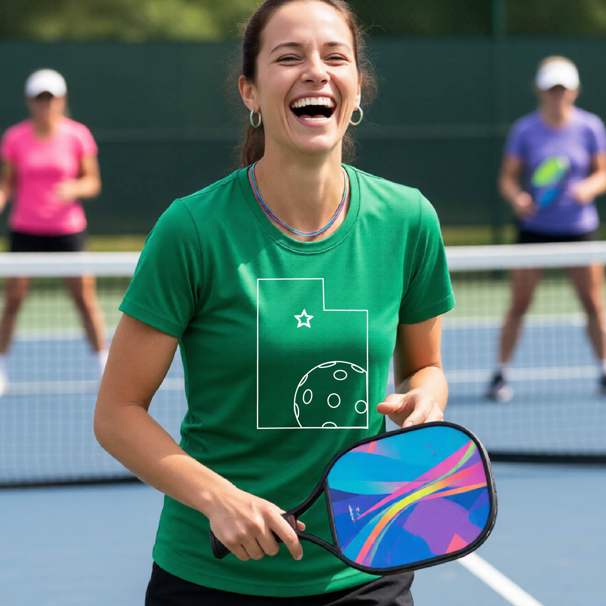 A close-up of a woman wearing a green short-sleeve, with a white graphic outline of the State of Utah , including an outline of a white pickleball in the lower right corner and a star outline in the center of the graphic, on the back of the shirt.   