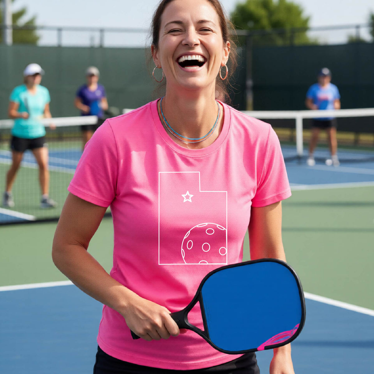 A close-up of a woman wearing a pink short-sleeve, with a white graphic outline of the State of Utah , including an outline of a white pickleball in the lower right corner and a star outline in the center of the graphic, on the back of the shirt.   