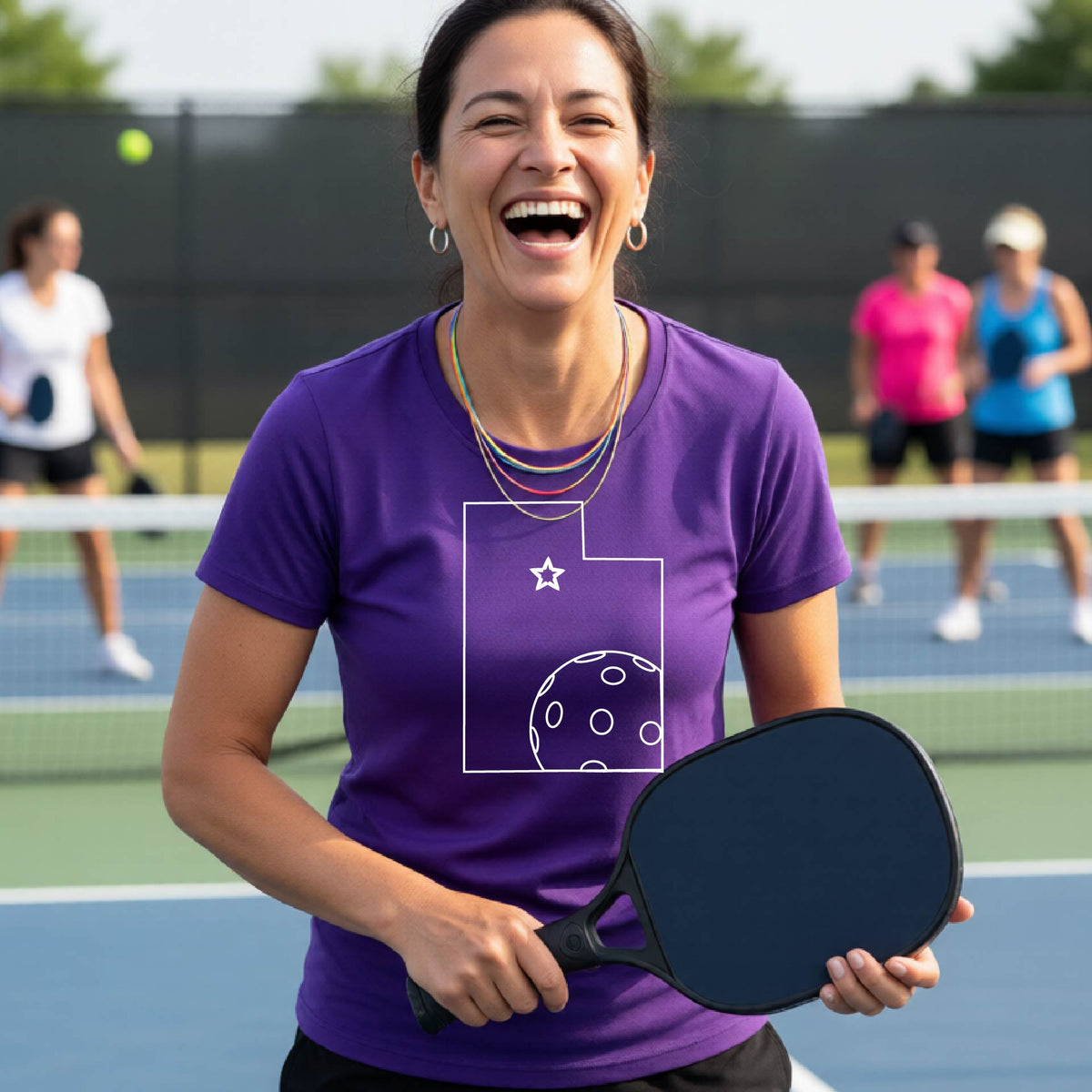 A close-up of a woman wearing a purple short-sleeve, with a white graphic outline of the State of Utah , including an outline of a white pickleball in the lower right corner and a star outline in the center of the graphic, on the back of the shirt.   