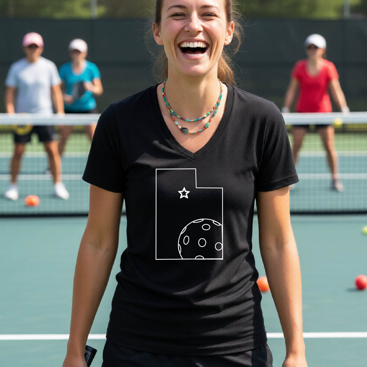 A close-up of a woman wearing a black long-sleeve, with a white graphic outline of the State of Utah , including an outline of a white pickleball in the lower right corner and a star outline in the center of the graphic, on the back of the shirt.   

