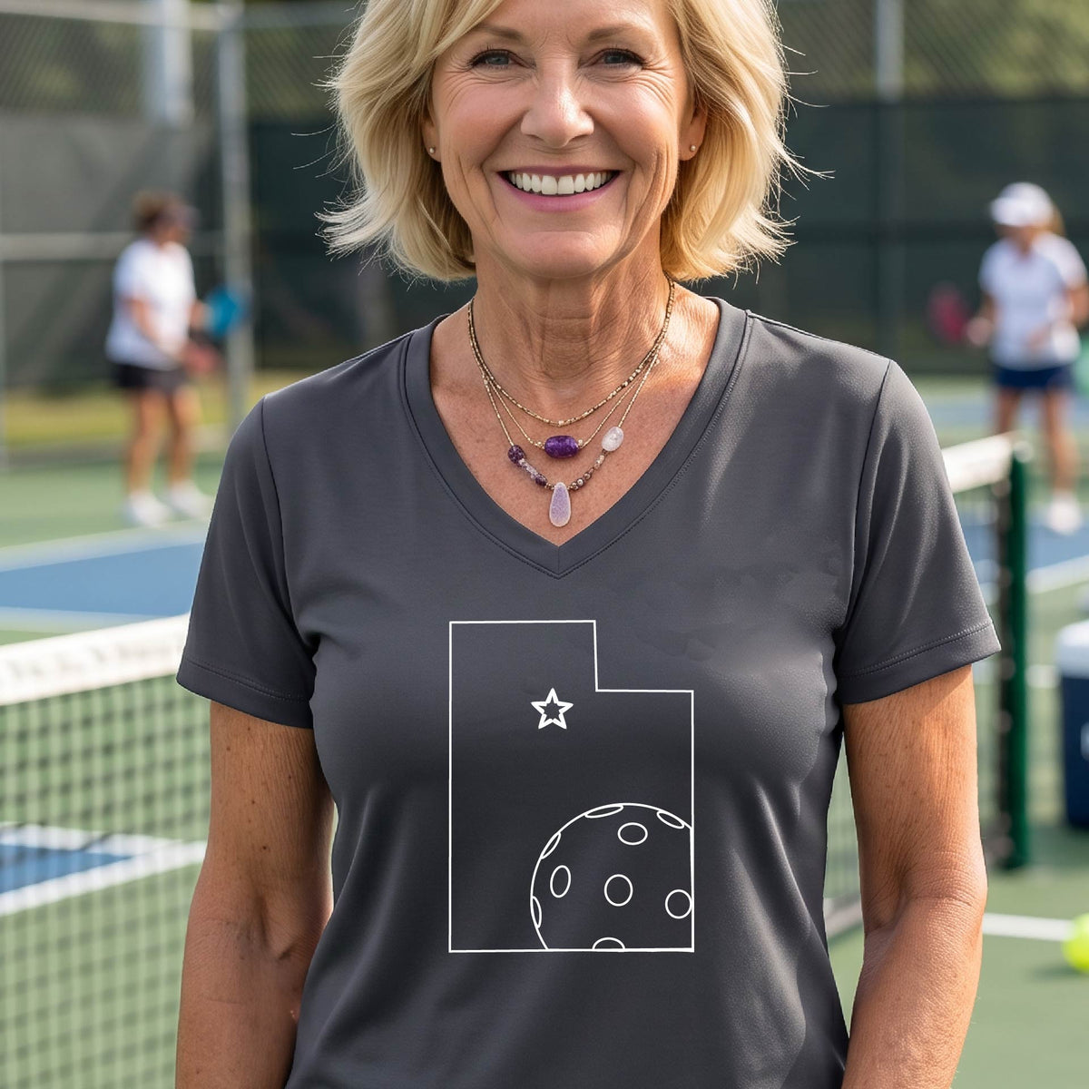A close-up of a woman wearing a dark gray long-sleeve, with a white graphic outline of the State of Utah , including an outline of a white pickleball in the lower right corner and a star outline in the center of the graphic, on the back of the shirt.   