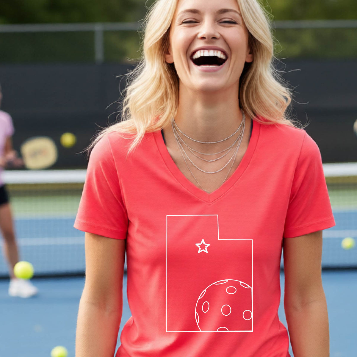 A close-up of a woman wearing a coral pink long-sleeve, with a white graphic outline of the State of Utah , including an outline of a white pickleball in the lower right corner and a star outline in the center of the graphic, on the back of the shirt.   

