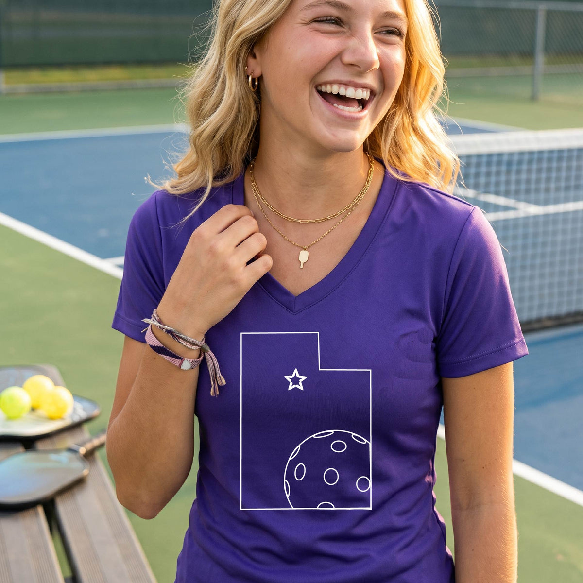 A close-up of a woman wearing a purple long-sleeve, with a white graphic outline of the State of Utah , including an outline of a white pickleball in the lower right corner and a star outline in the center of the graphic, on the back of the shirt.   

