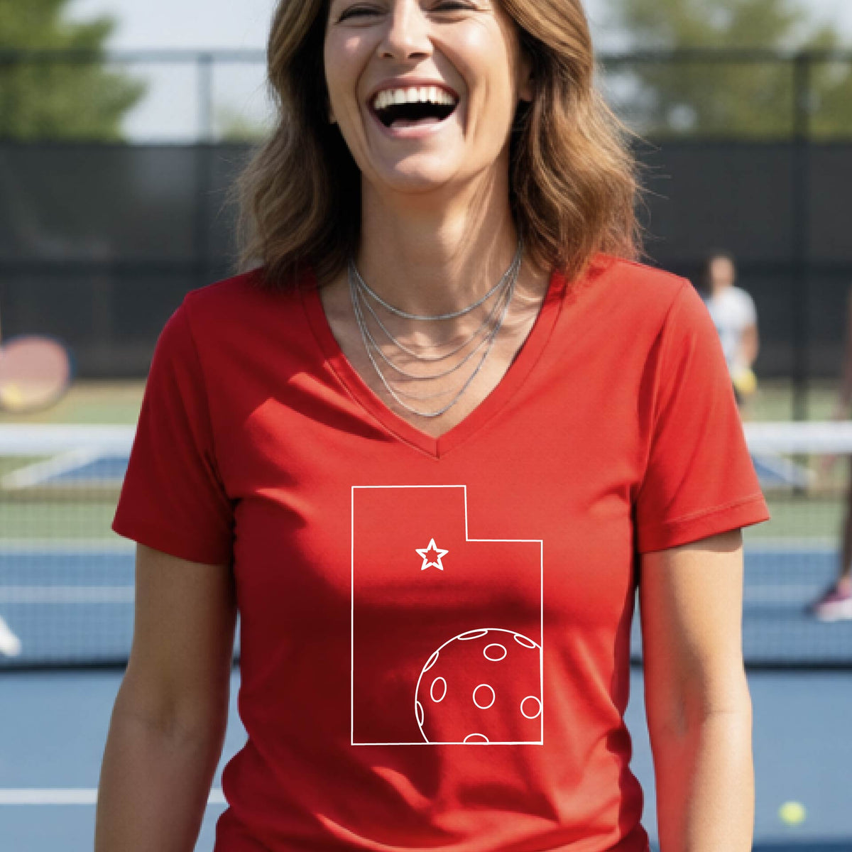 A close-up of a woman wearing a red long-sleeve, with a white graphic outline of the State of Utah , including an outline of a white pickleball in the lower right corner and a star outline in the center of the graphic, on the back of the shirt.   

