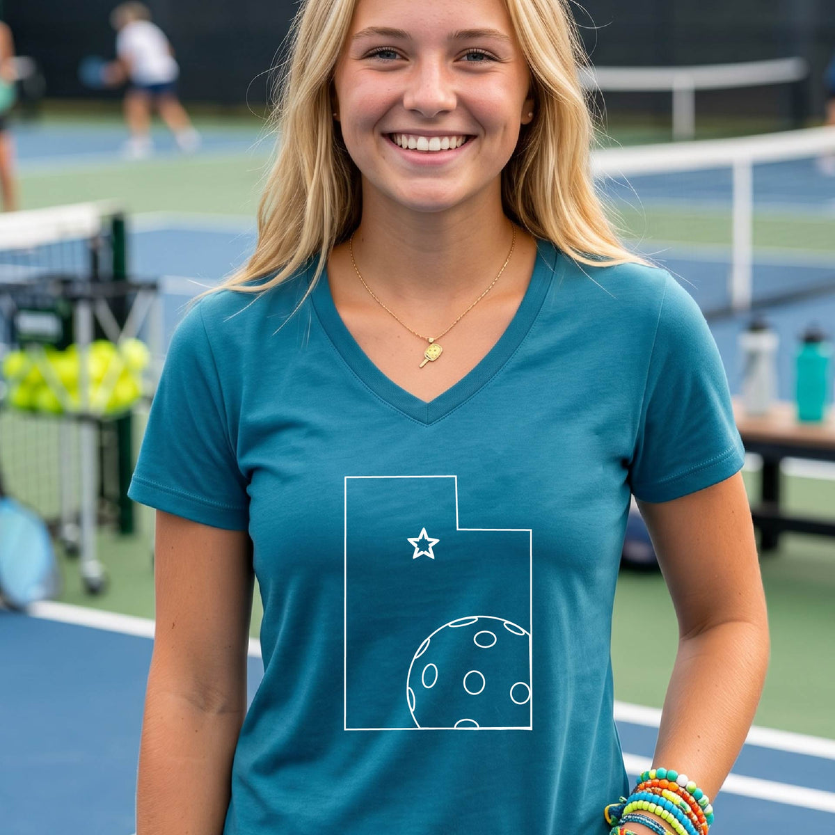 A close-up of a woman wearing a teal long-sleeve, with a white graphic outline of the State of Utah , including an outline of a white pickleball in the lower right corner and a star outline in the center of the graphic, on the back of the shirt.   

