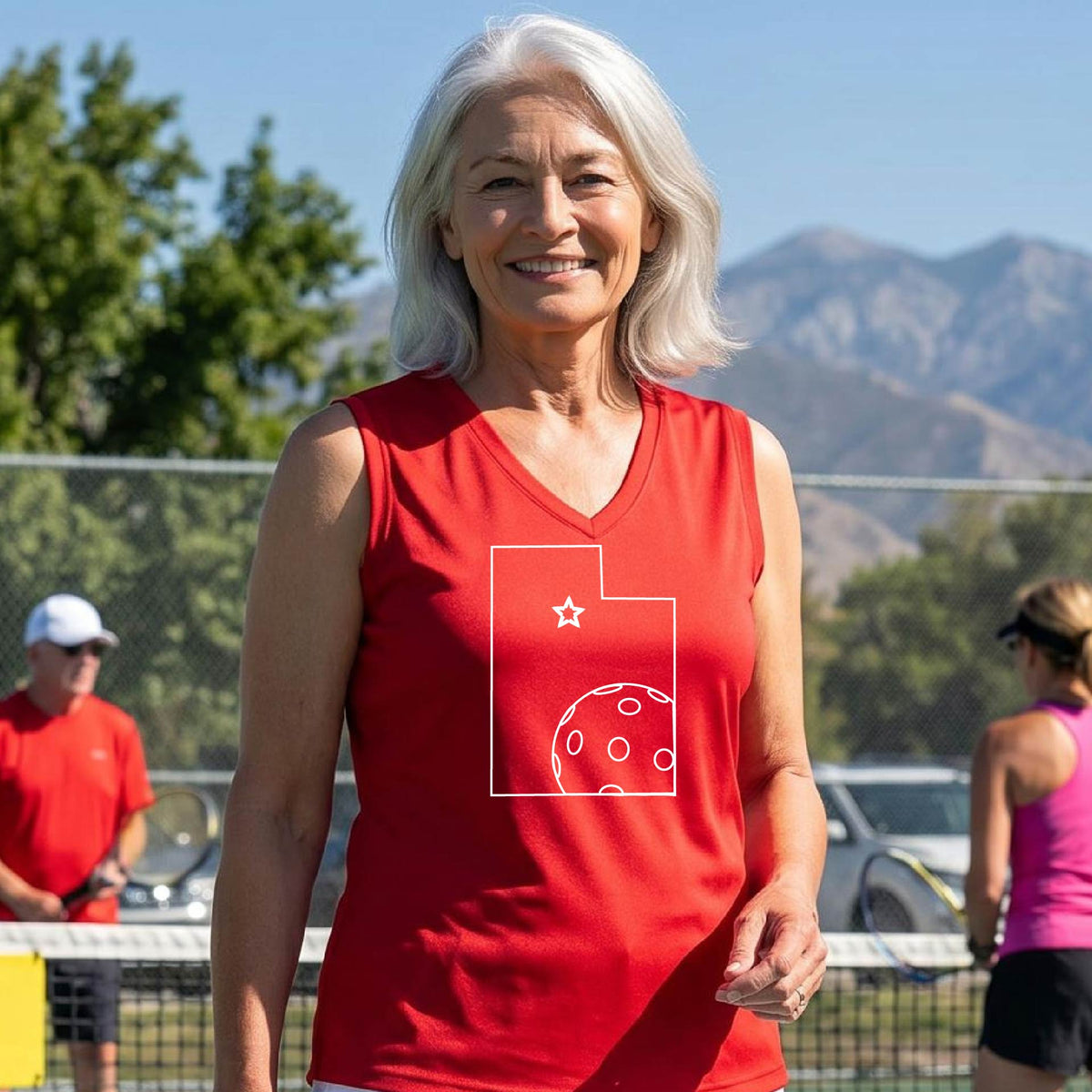 A close-up of a woman wearing a red sleeveless, with a white graphic outline of the State of Utah , including an outline of a white pickleball in the lower right corner and a star outline in the center of the graphic, on the back of the shirt. 