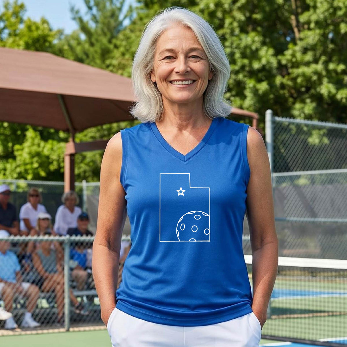A close-up of a woman wearing a royal blue sleeveless, with a white graphic outline of the State of Utah , including an outline of a white pickleball in the lower right corner and a star outline in the center of the graphic, on the back of the shirt. 