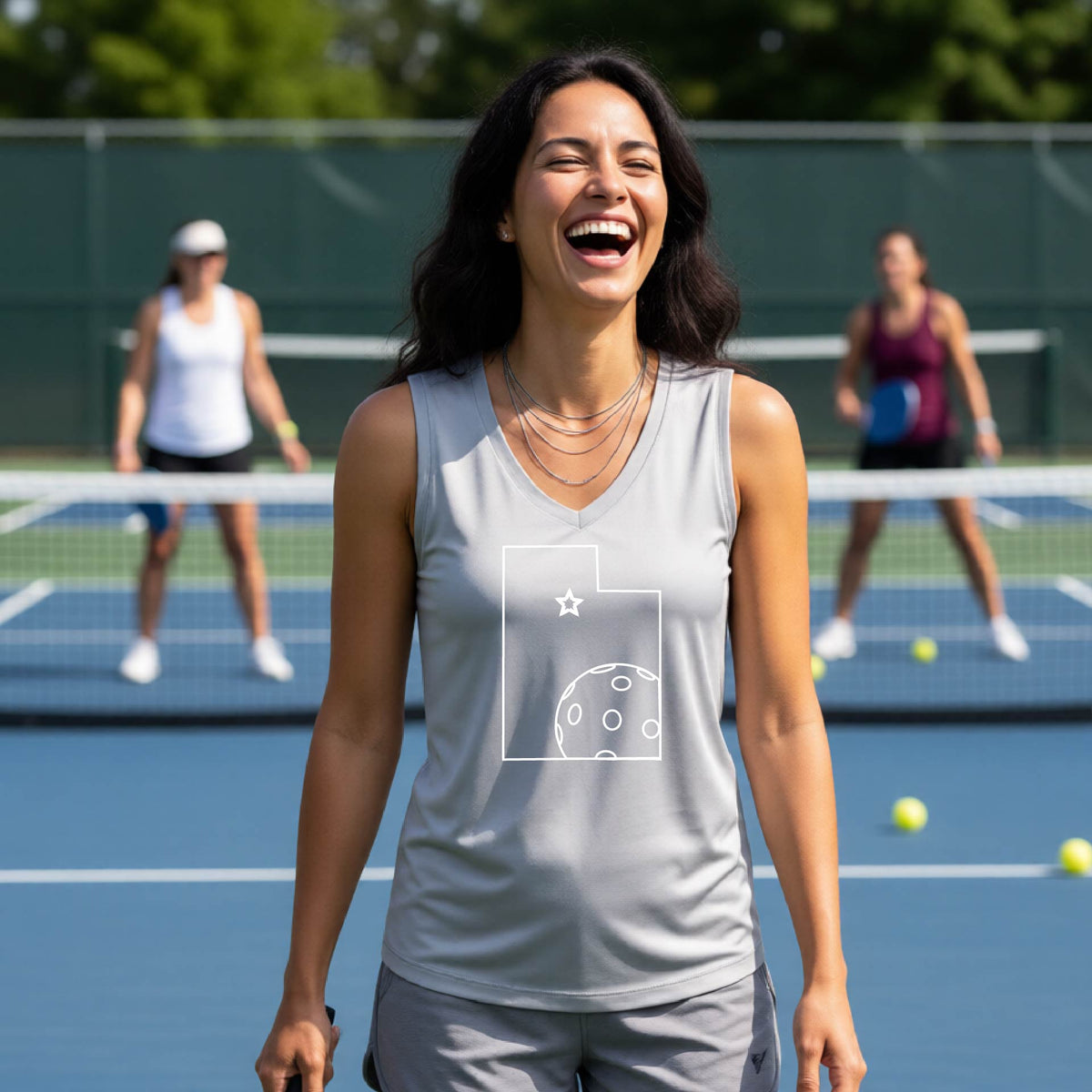 A close-up of a woman wearing a silver gray sleeveless, with a white graphic outline of the State of Utah , including an outline of a white pickleball in the lower right corner and a star outline in the center of the graphic, on the back of the shirt. 