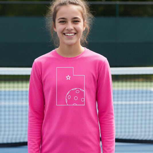 A close-up of a young person wearing a pink long-sleeve t-shirt, with a white graphic of the State of Utah , including an outline of a white pickleball in the lower right corner and a star outline in the center of the graphic, on the front of the shirt.   