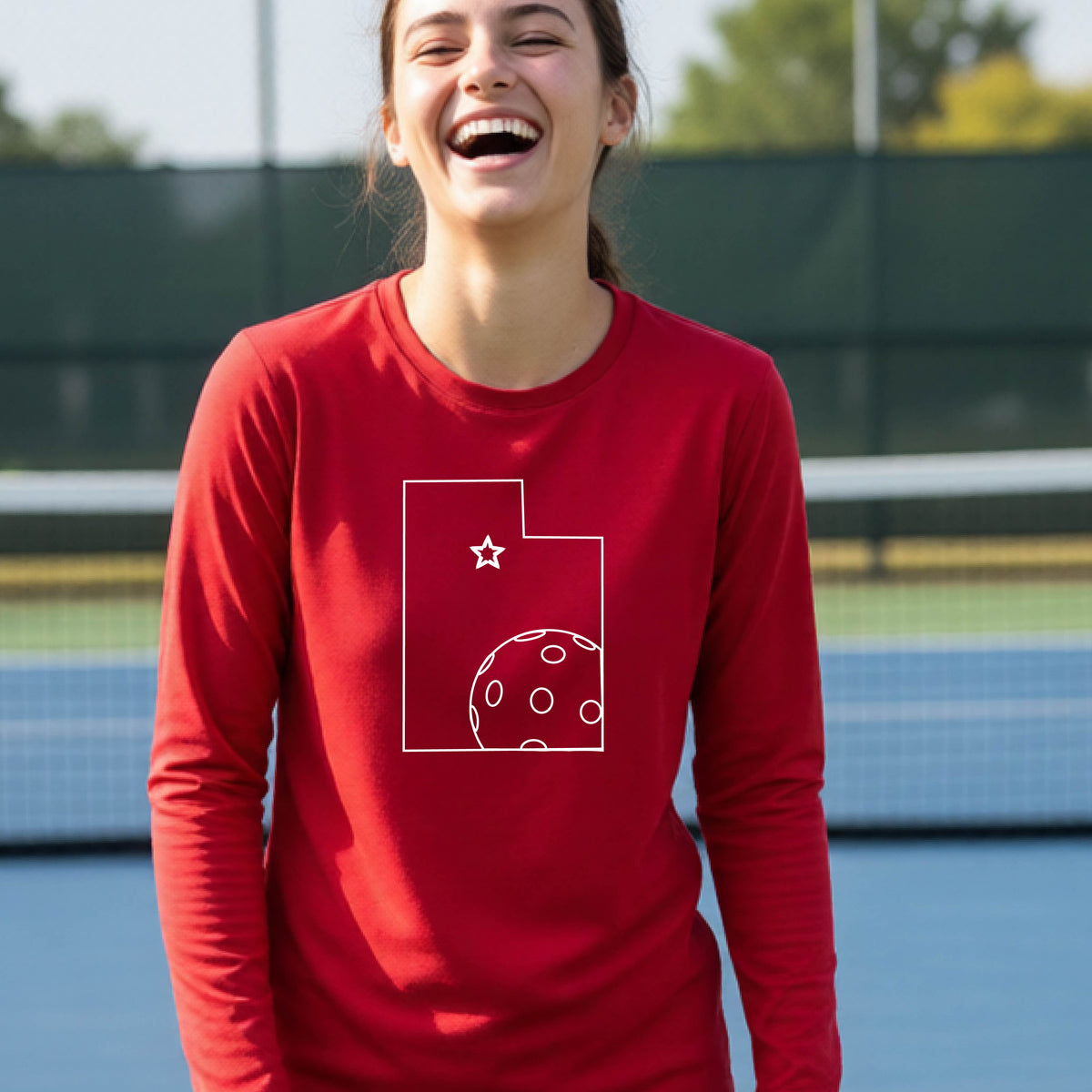 A close-up of a young person wearing a red long-sleeve t-shirt, with a white graphic of the State of Utah , including an outline of a white pickleball in the lower right corner and a star outline in the center of the graphic, on the front of the shirt.   