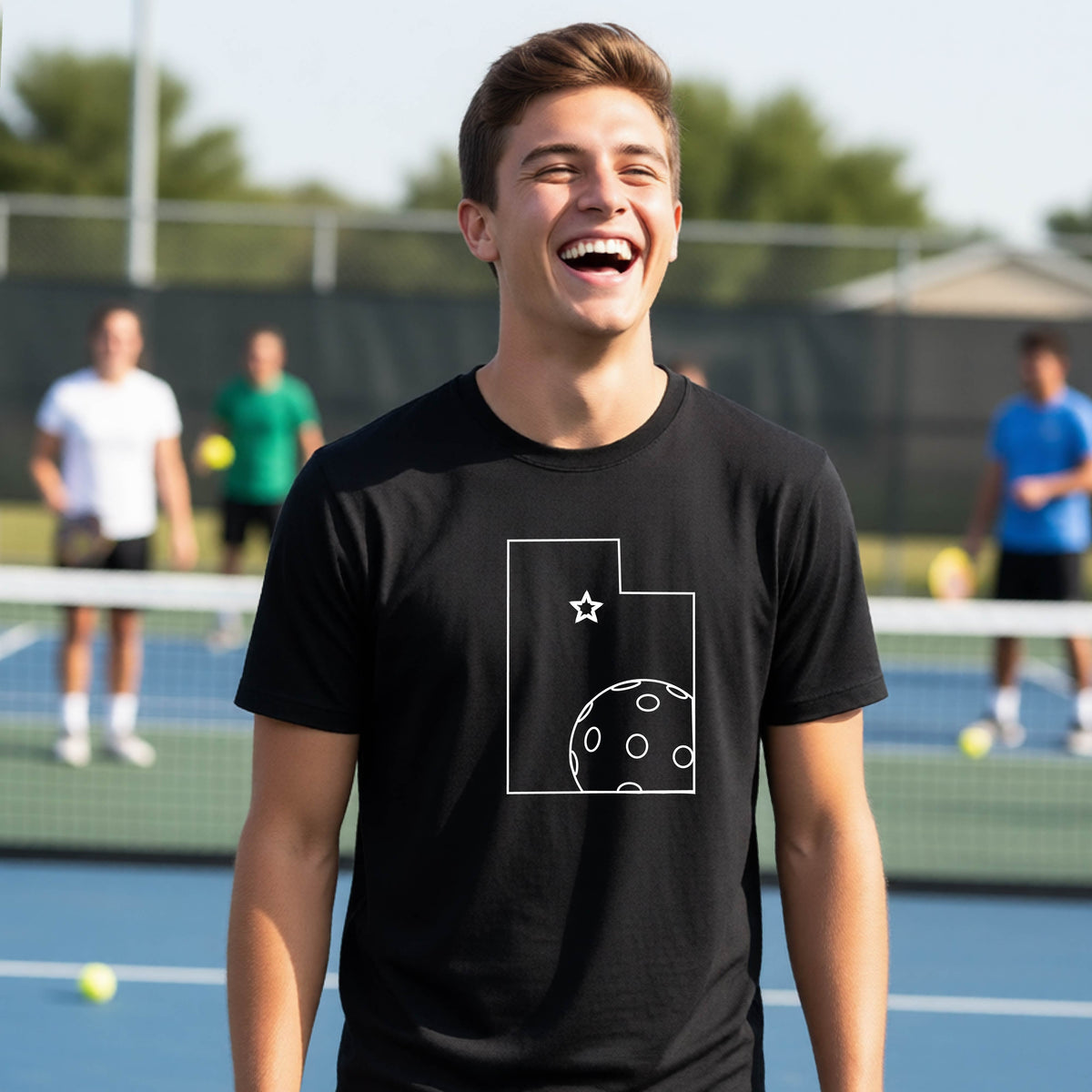 A close-up of a young person wearing a black short-sleeve shirt, with a white graphic of the State of Utah , including an outline of a white pickleball in the lower right corner and a star outline in the center of the graphic, on the front of the shirt.   