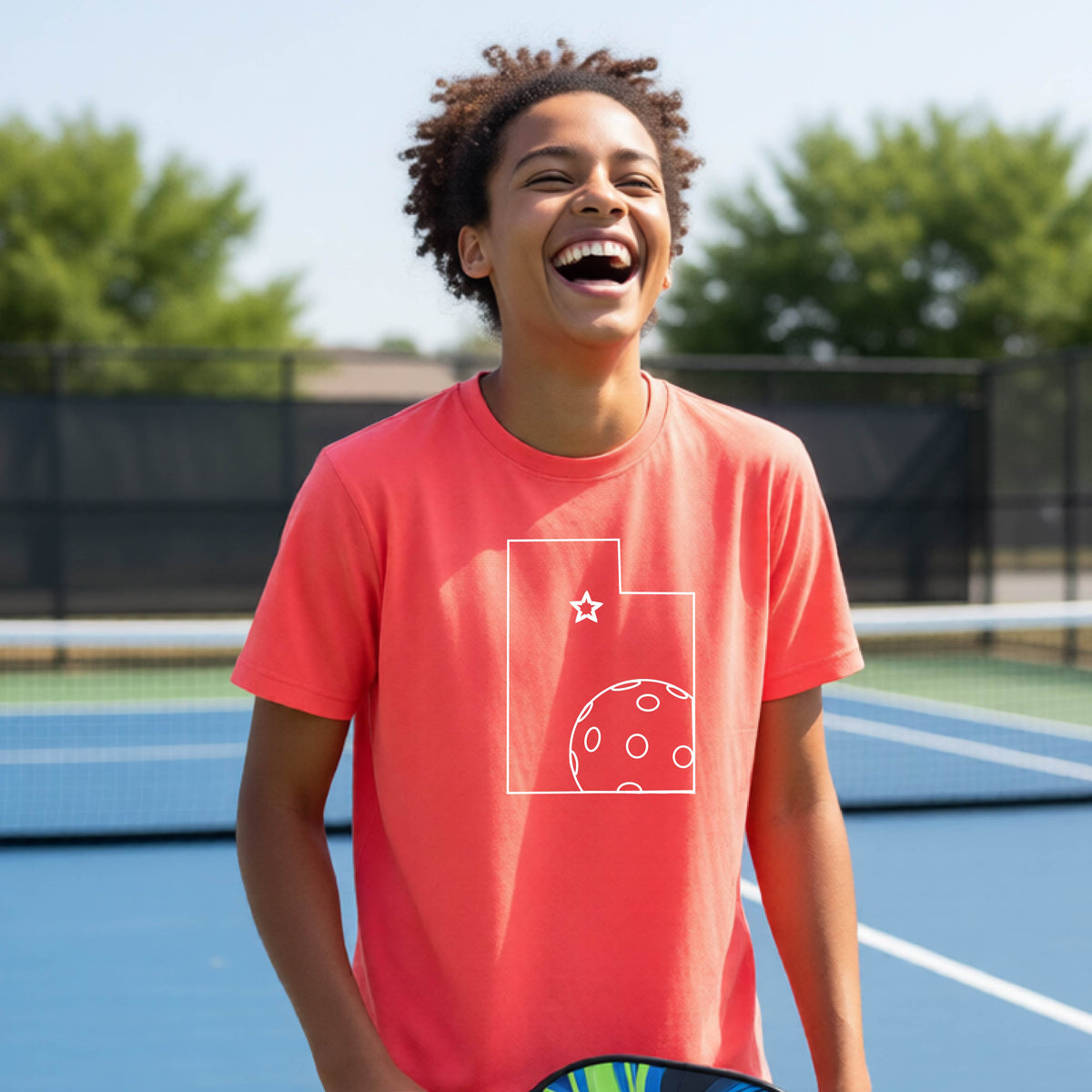A close-up of a young person wearing a coral pink short-sleeve shirt, with a white graphic of the State of Utah , including an outline of a white pickleball in the lower right corner and a star outline in the center of the graphic, on the front of the shirt.   