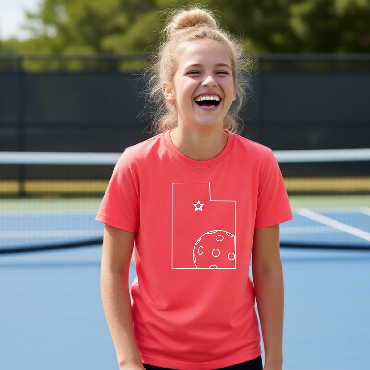 A close-up of a young person wearing a coral pink short-sleeve shirt, with a white graphic of the State of Utah , including an outline of a white pickleball in the lower right corner and a star outline in the center of the graphic, on the front of the shirt.   