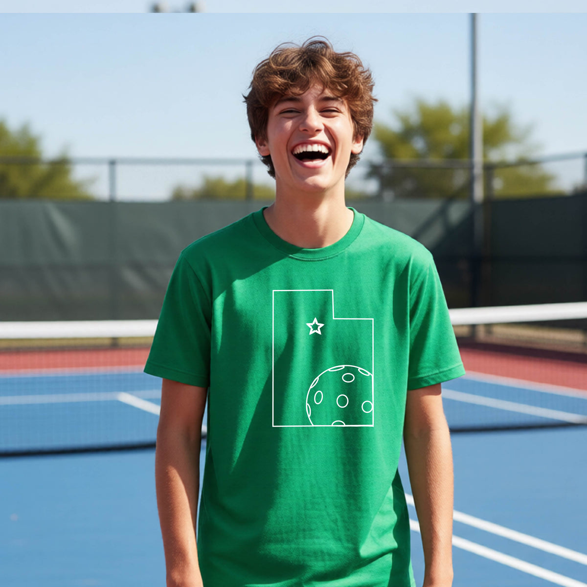 A close-up of a young person wearing a green short-sleeve shirt, with a white graphic of the State of Utah , including an outline of a white pickleball in the lower right corner and a star outline in the center of the graphic, on the front of the shirt.   