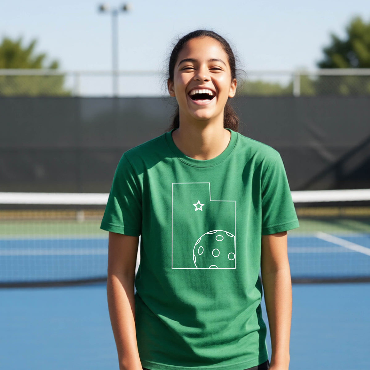 A close-up of a young person wearing a green short-sleeve shirt, with a white graphic of the State of Utah , including an outline of a white pickleball in the lower right corner and a star outline in the center of the graphic, on the front of the shirt.   