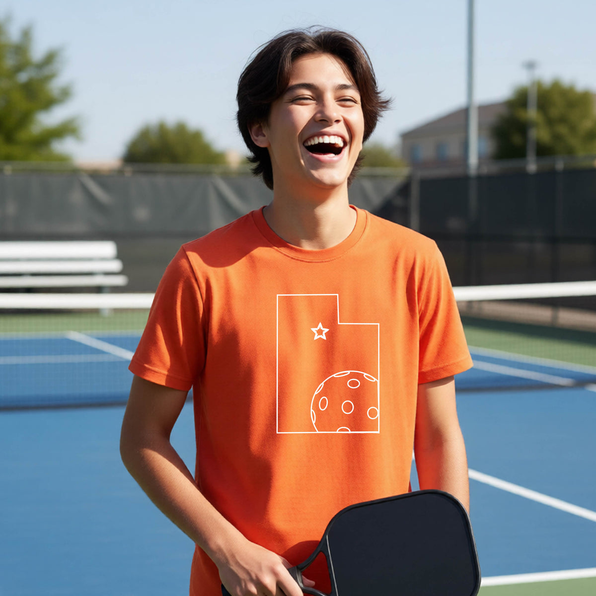 A close-up of a young person wearing an orange short-sleeve shirt, with a white graphic of the State of Utah , including an outline of a white pickleball in the lower right corner and a star outline in the center of the graphic, on the front of the shirt.   
