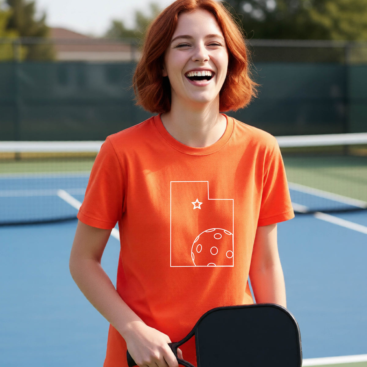 A close-up of a young person wearing an orange short-sleeve shirt, with a white graphic of the State of Utah , including an outline of a white pickleball in the lower right corner and a star outline in the center of the graphic, on the front of the shirt.   