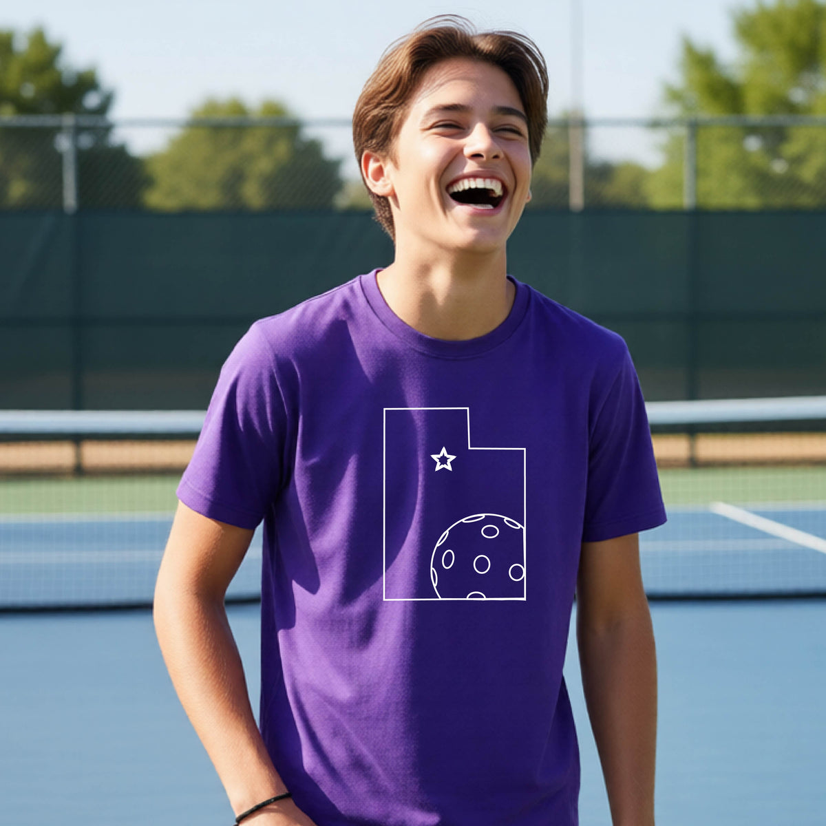 A close-up of a young person wearing a purple short-sleeve shirt, with a white graphic of the State of Utah , including an outline of a white pickleball in the lower right corner and a star outline in the center of the graphic, on the front of the shirt.   