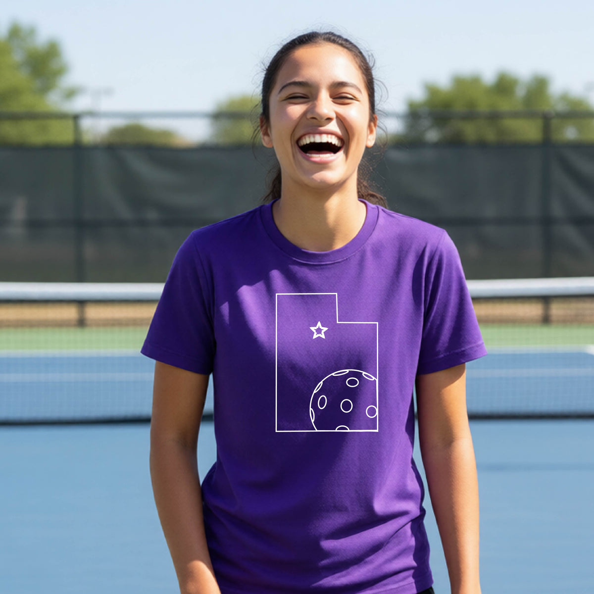 A close-up of a young person wearing a purple short-sleeve shirt, with a white graphic of the State of Utah , including an outline of a white pickleball in the lower right corner and a star outline in the center of the graphic, on the front of the shirt.   