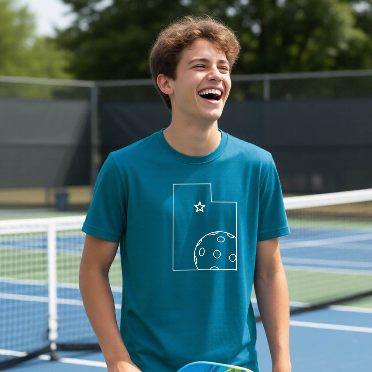 A close-up of a young person wearing a teal short-sleeve shirt, with a white graphic of the State of Utah , including an outline of a white pickleball in the lower right corner and a star outline in the center of the graphic, on the front of the shirt.   