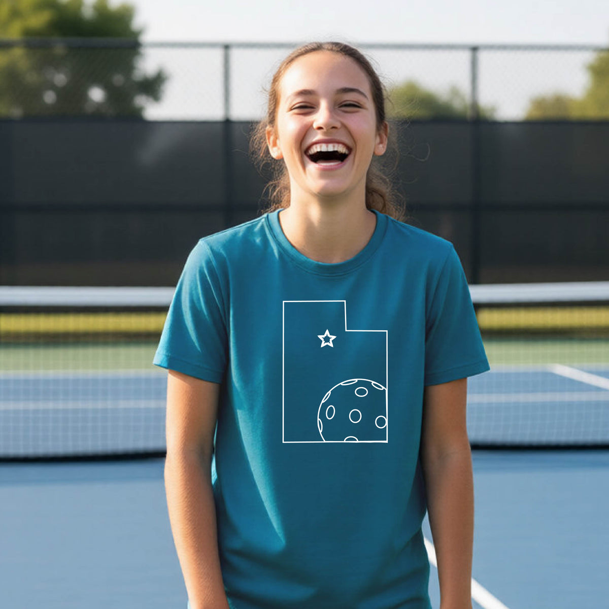 A close-up of a young person wearing a teal short-sleeve shirt, with a white graphic of the State of Utah , including an outline of a white pickleball in the lower right corner and a star outline in the center of the graphic, on the front of the shirt.    