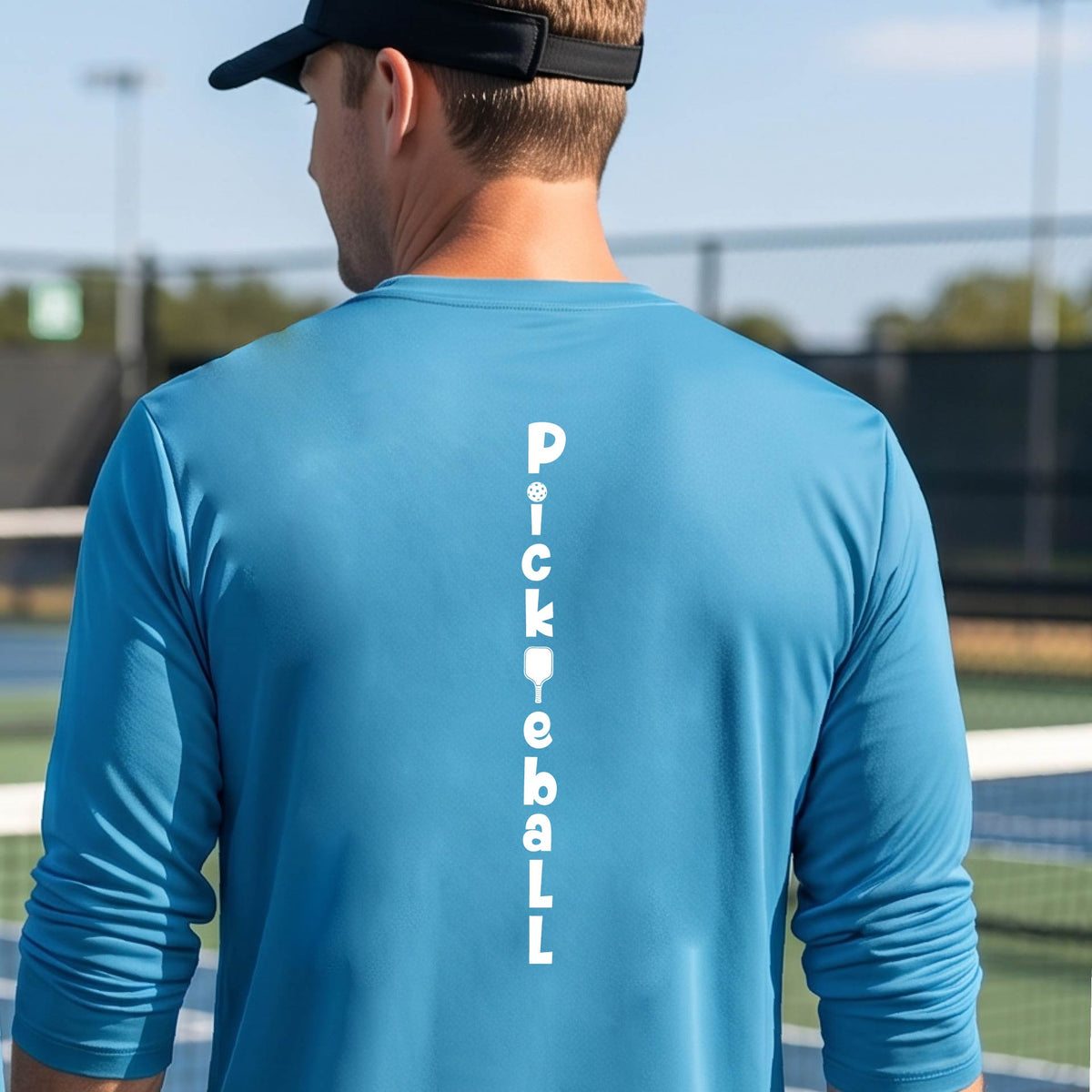 A man wearing a bright blue long-sleeve t-shirt with a white graphic text 'PICKLEBALL' on the back with a pickleball paddle replacing the L, standing on a pickleball court.