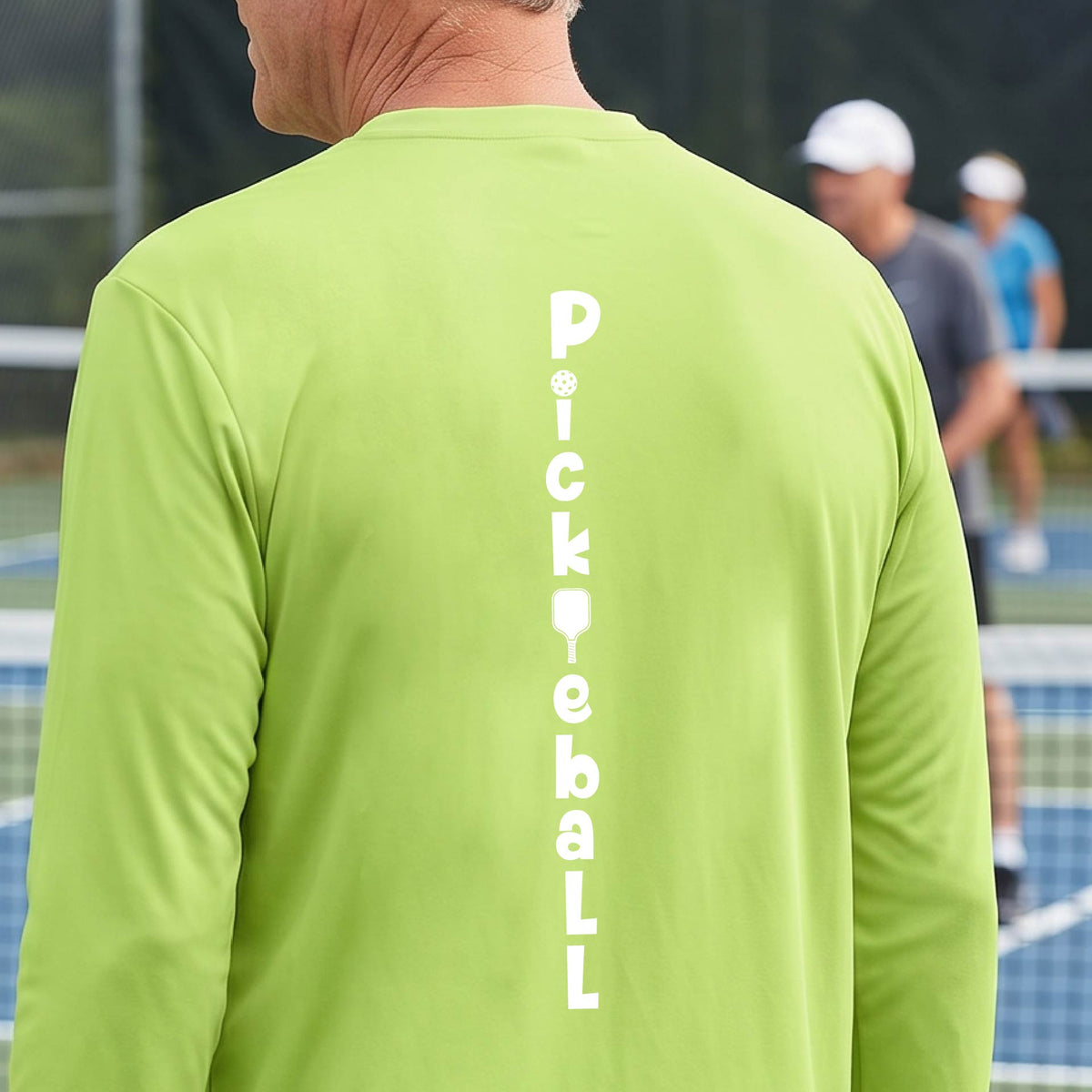 A man wearing a lime green long-sleeve t-shirt with a white graphic text 'PICKLEBALL' on the back with a pickleball paddle replacing the L, standing on a pickleball court.