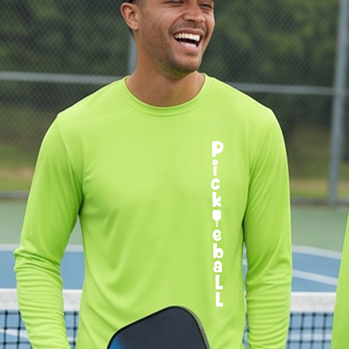 A man wearing a lime green long-sleeve t-shirt with a white graphic text 'PICKLEBALL' on the front with a pickleball paddle replacing the L, standing on a pickleball court.