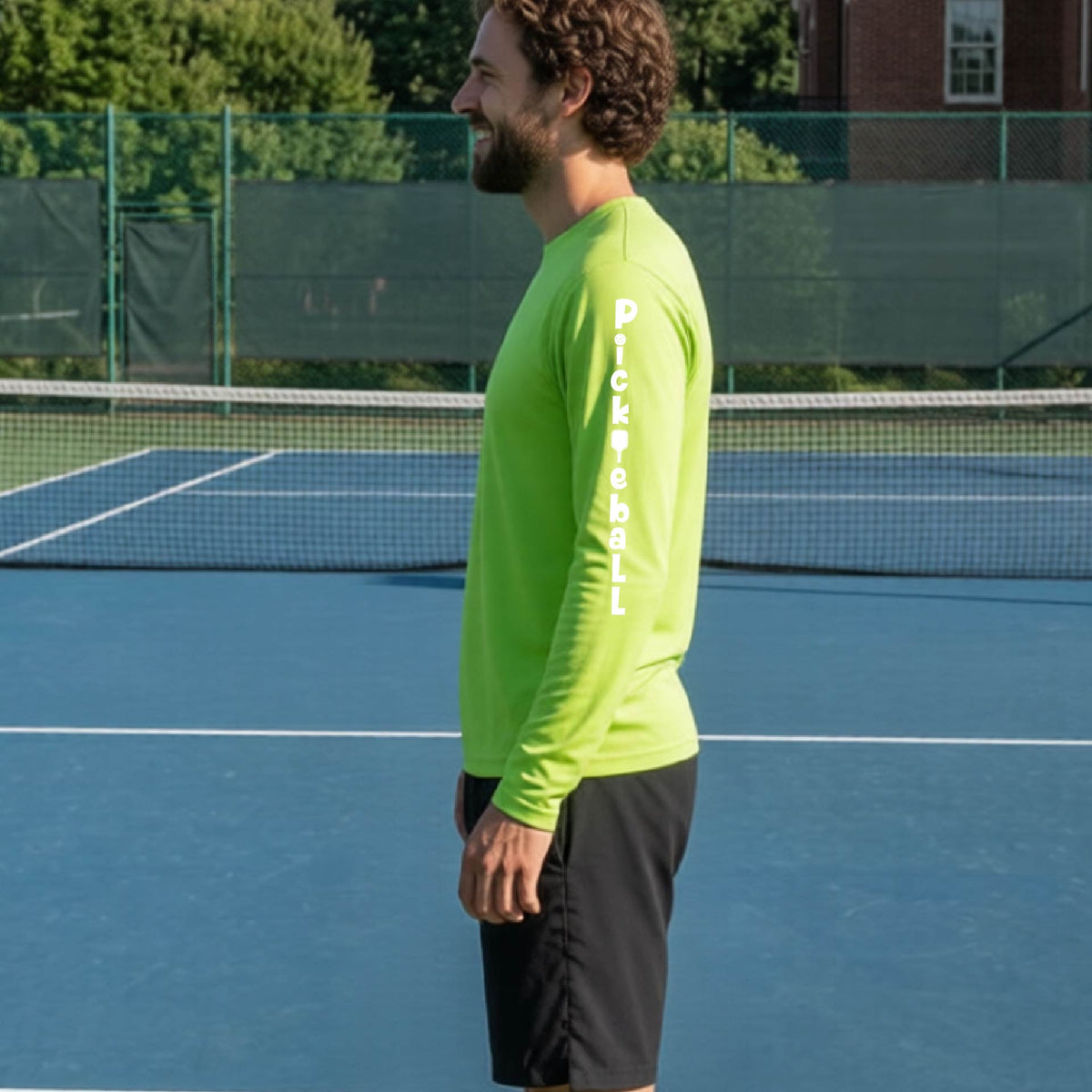 A man wearing a lime green long-sleeve t-shirt with a white graphic text 'PICKLEBALL' on the sleeve with a pickleball paddle replacing the L, standing on a pickleball court.