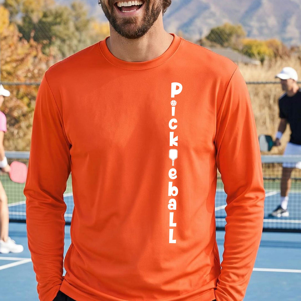 A man wearing an orange long-sleeve t-shirt with a white graphic text 'PICKLEBALL' on the front with a pickleball paddle replacing the L, standing on a pickleball court.
