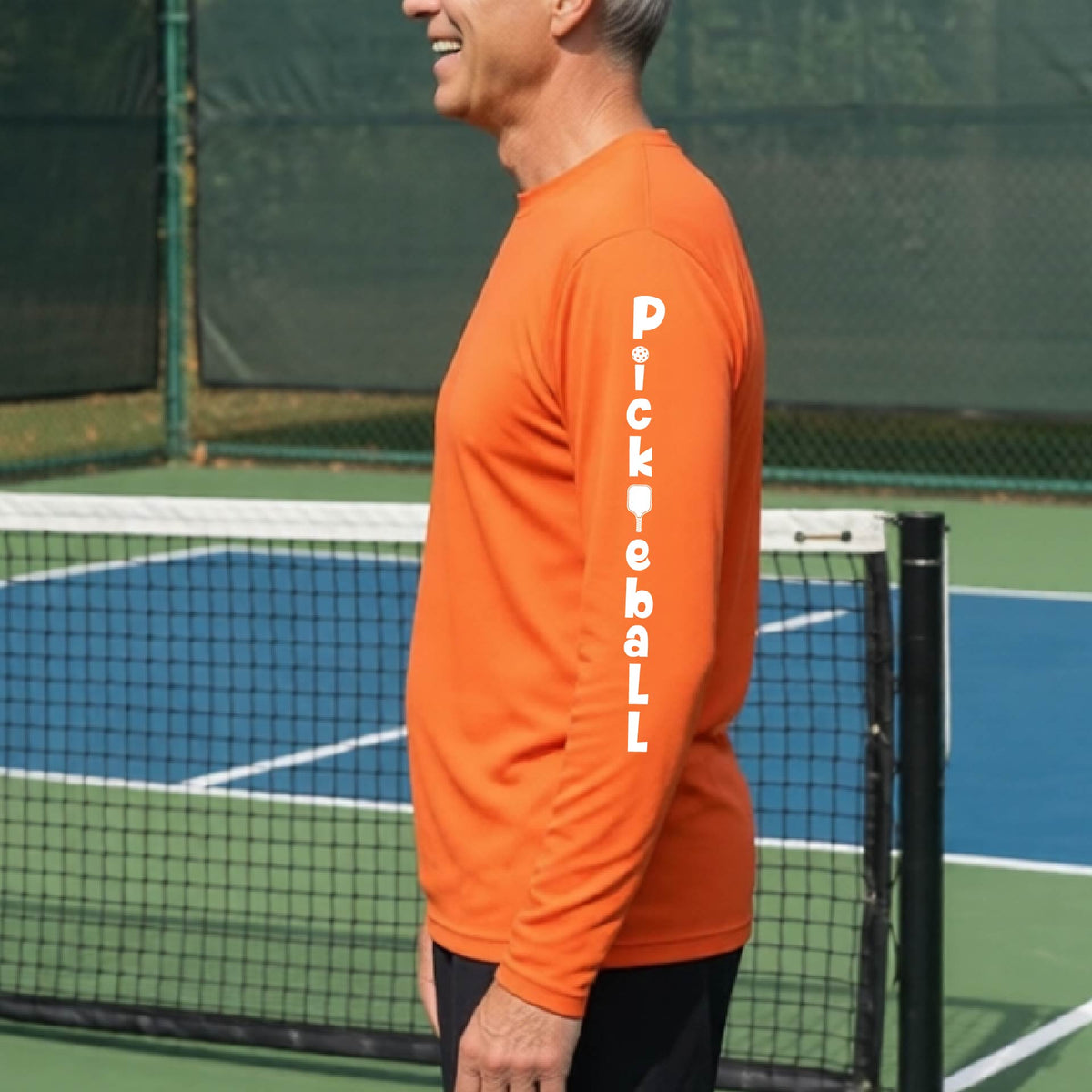 A man wearing an orange long-sleeve t-shirt with a white graphic text 'PICKLEBALL' on the sleeve with a pickleball paddle replacing the L, standing on a pickleball court.