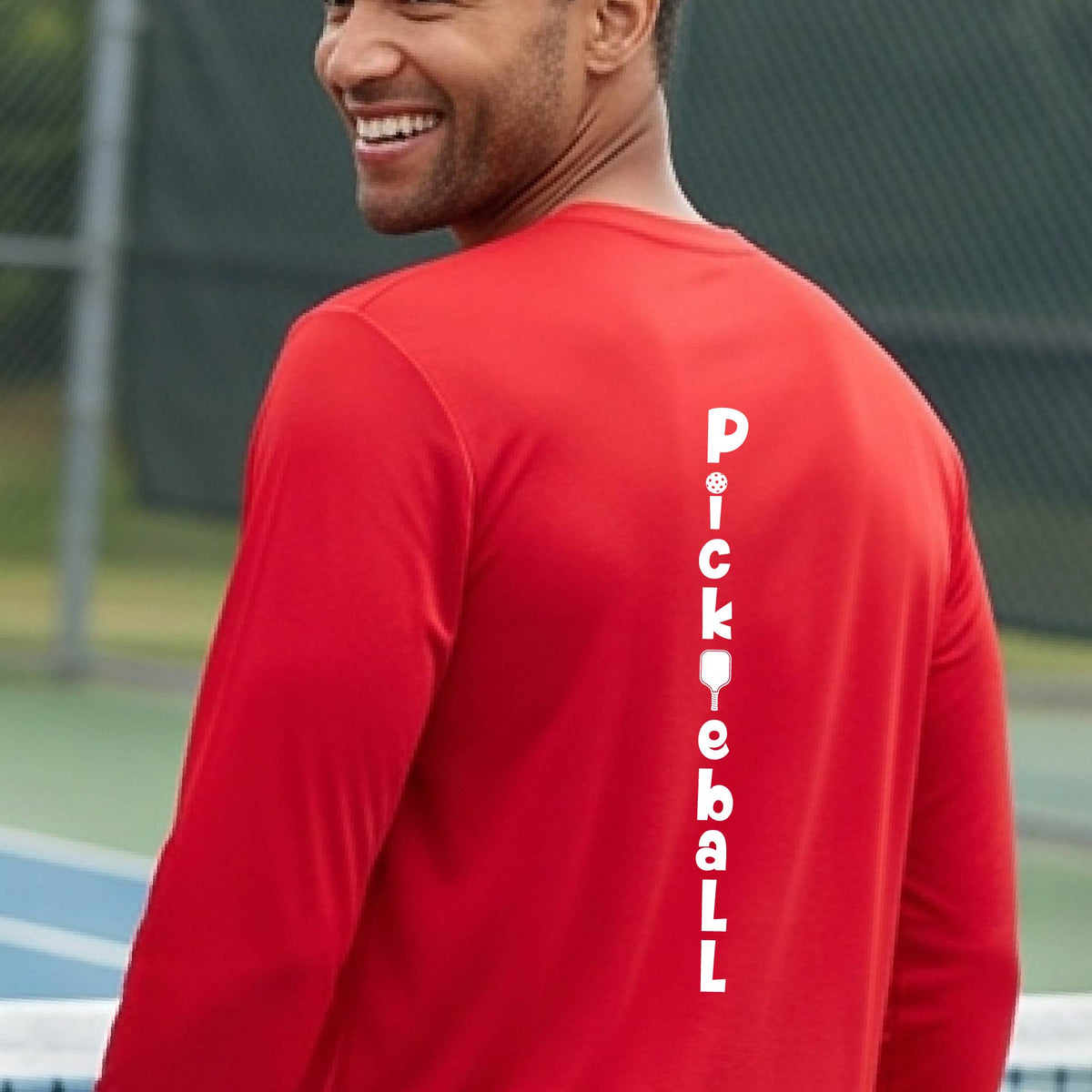 A man wearing a red long-sleeve t-shirt with a white graphic text 'PICKLEBALL' on the back with a pickleball paddle replacing the L, standing on a pickleball court.
