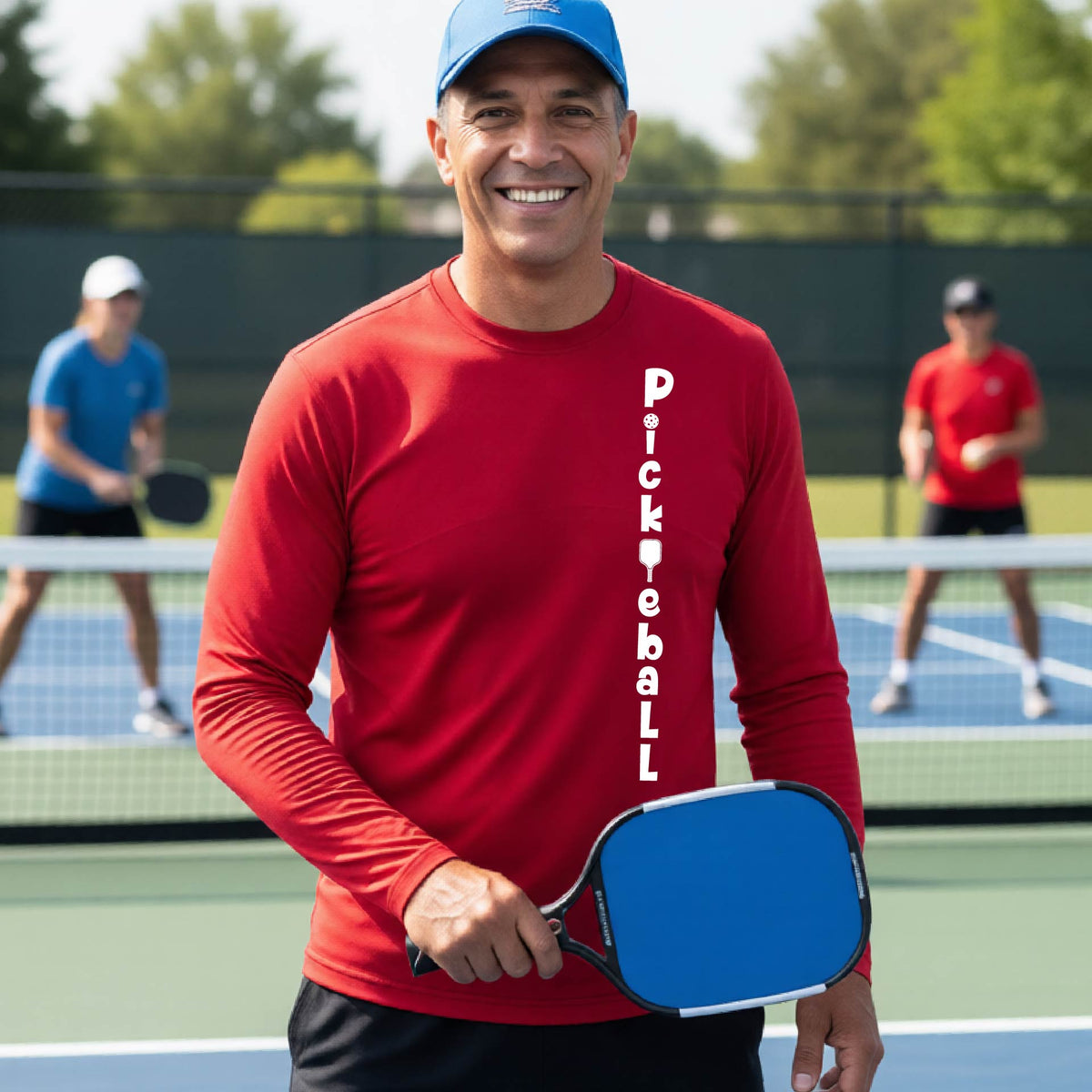 A man wearing a red long-sleeve t-shirt with a white graphic text 'PICKLEBALL' on the front with a pickleball paddle replacing the L, standing on a pickleball court.