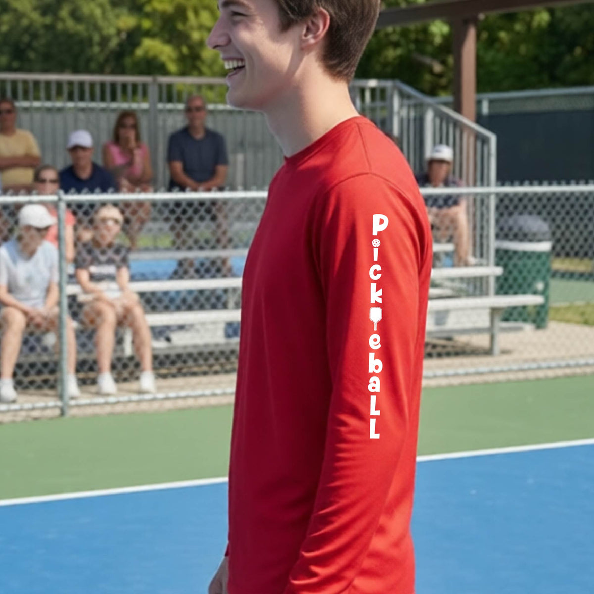 A man wearing a red long-sleeve t-shirt with a white graphic text 'PICKLEBALL' on the sleeve with a pickleball paddle replacing the L, standing on a pickleball court.