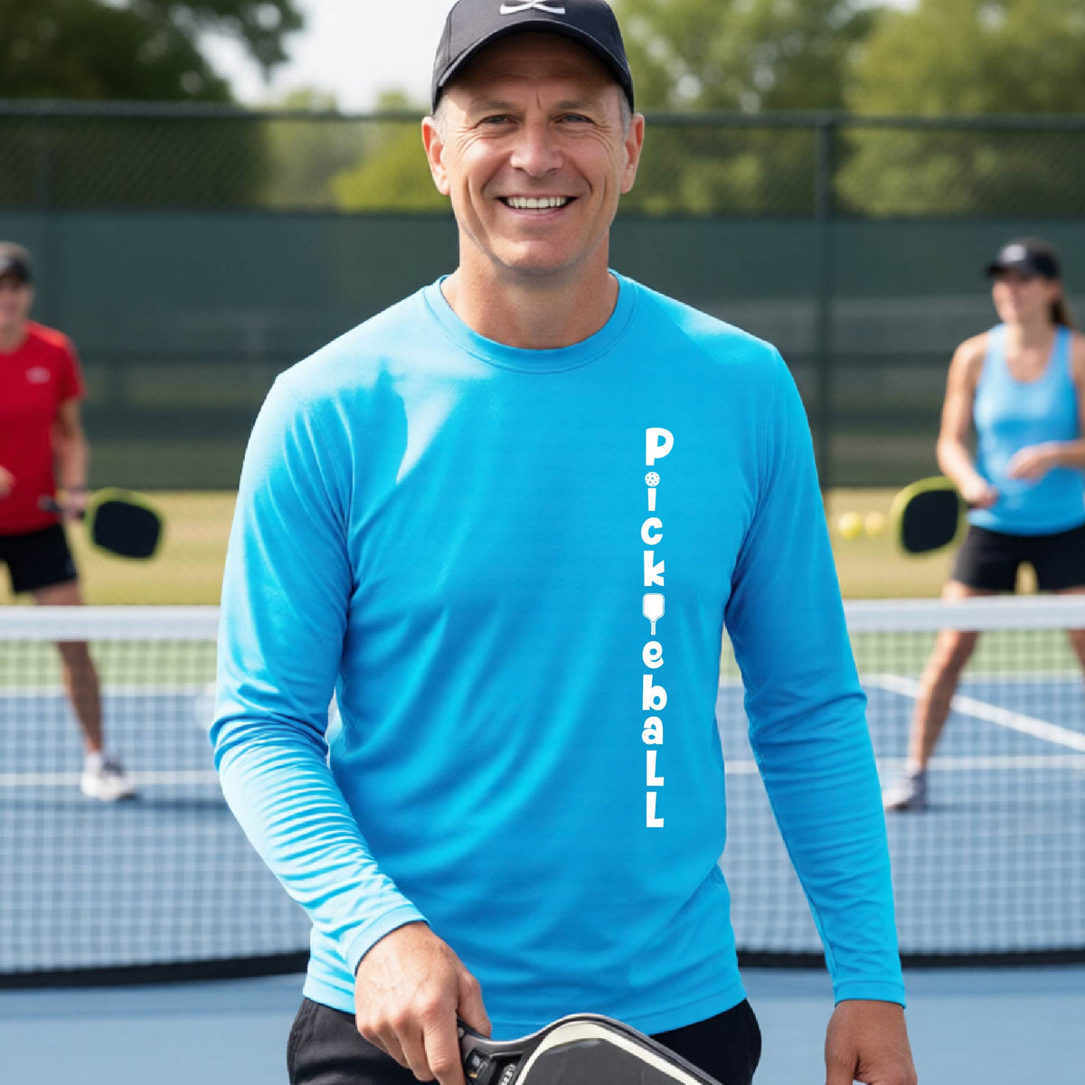 A man wearing a bright blue long-sleeve t-shirt with a white graphic text 'PICKLEBALL' on the front with a pickleball paddle replacing the L, standing on a pickleball court.
