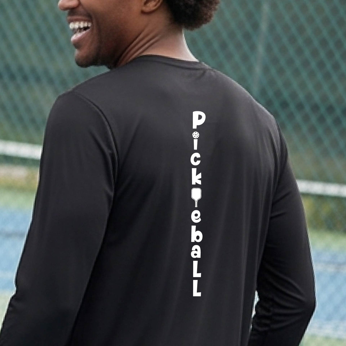 A man wearing a black long-sleeve t-shirt with a white graphic text 'PICKLEBALL' on the back with a pickleball paddle replacing the L, standing on a pickleball court.