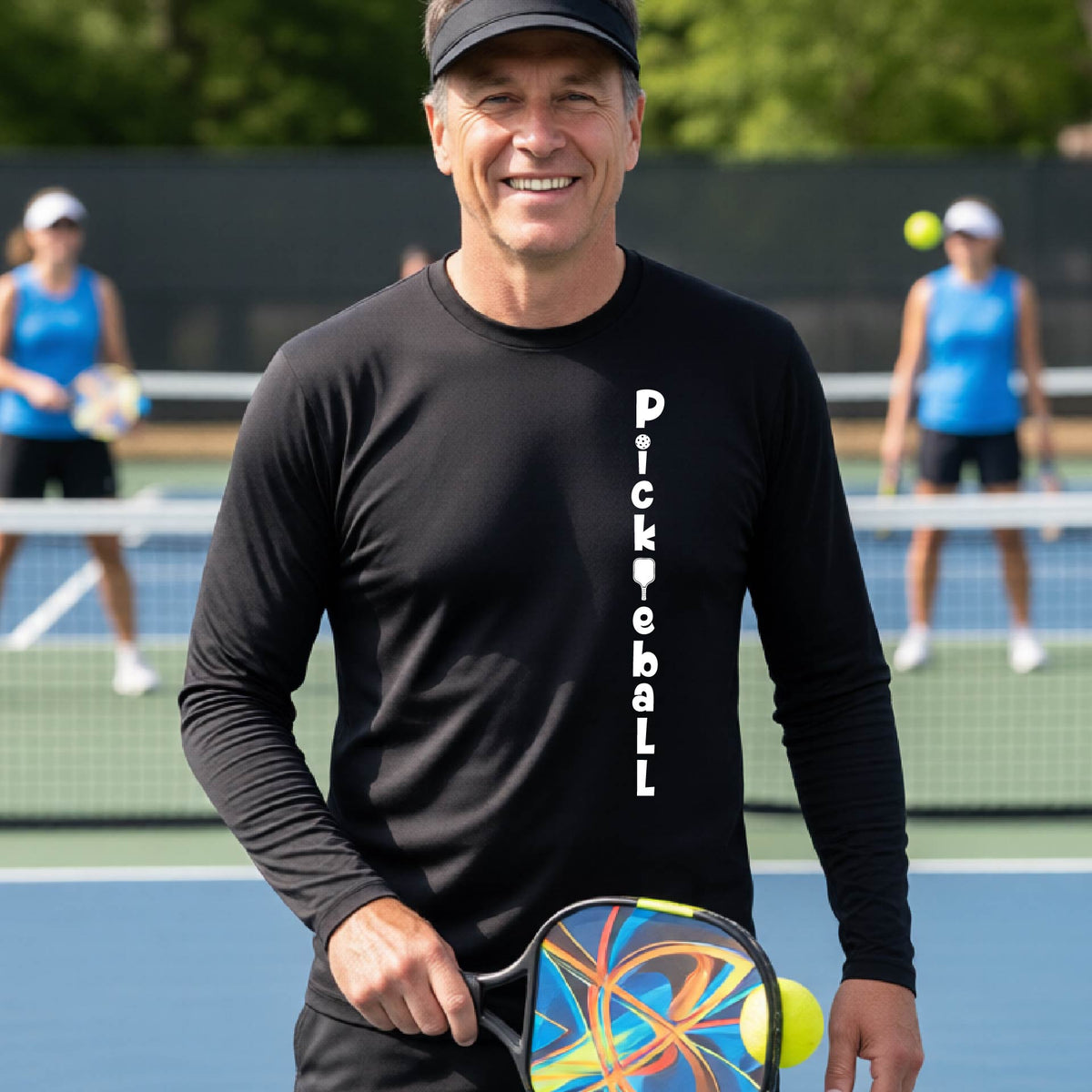 A man wearing a black long-sleeve t-shirt with a white graphic text 'PICKLEBALL' on the front with a pickleball paddle replacing the L, standing on a pickleball court.