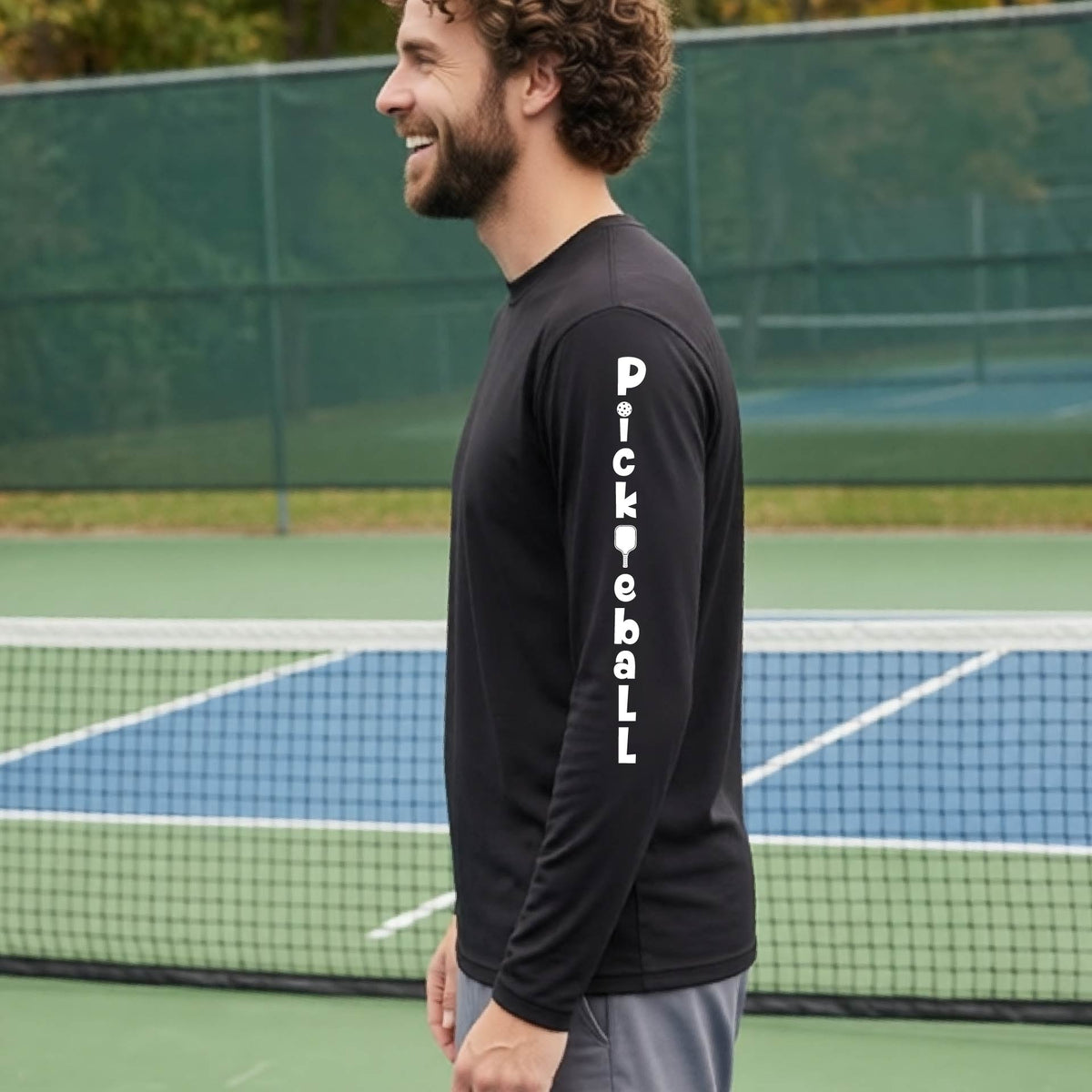 A man wearing a black long-sleeve t-shirt with a white graphic text 'PICKLEBALL' on the sleeve with a pickleball paddle replacing the L, standing on a pickleball court.