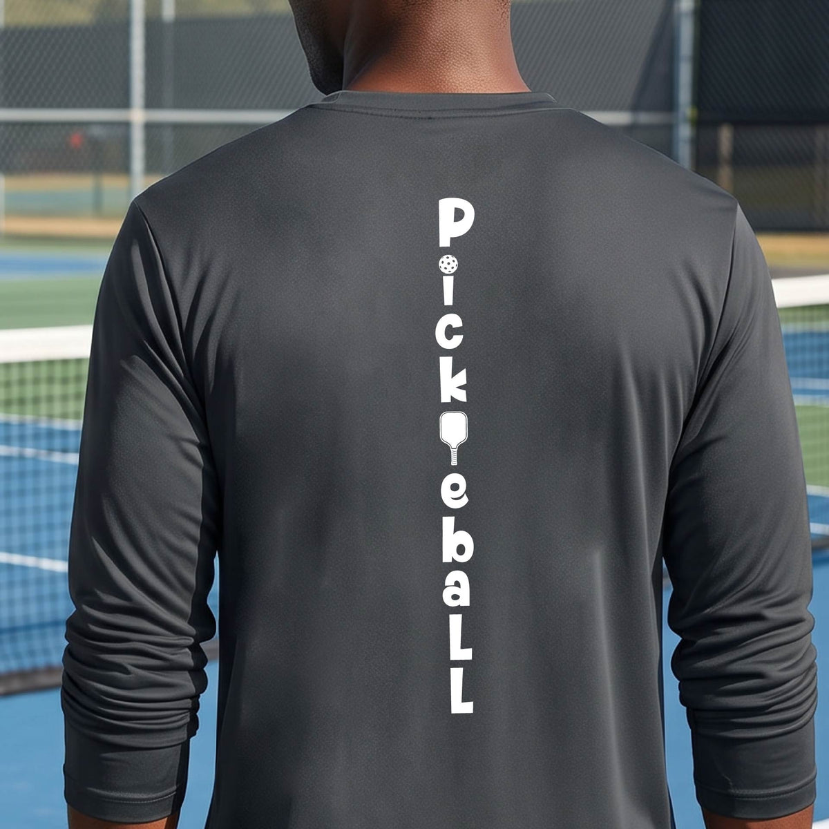 A man wearing a gray long-sleeve t-shirt with a white graphic text 'PICKLEBALL' on the back with a pickleball paddle replacing the L, standing on a pickleball court.
