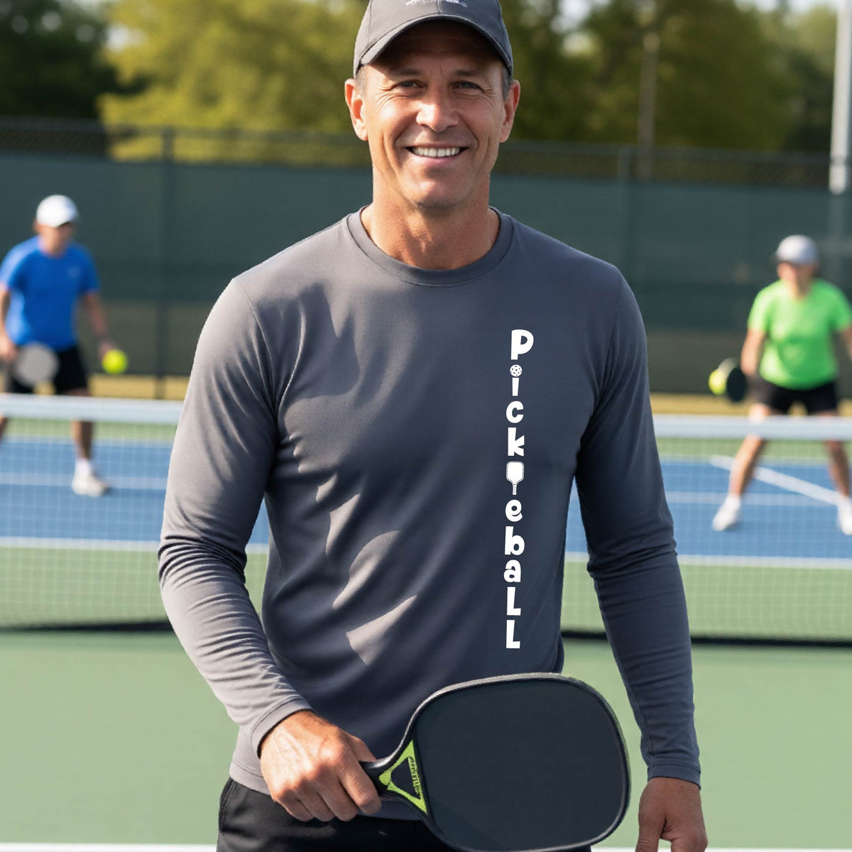 A man wearing a gray long-sleeve t-shirt with a white graphic text 'PICKLEBALL' on the front with a pickleball paddle replacing the L, standing on a pickleball court.