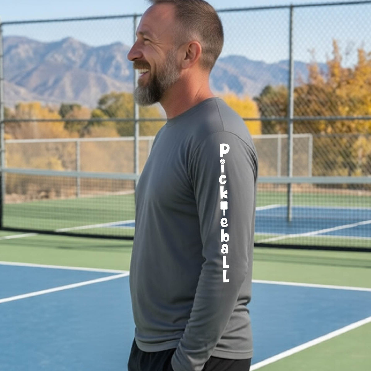 A man wearing a gray long-sleeve t-shirt with a white graphic text 'PICKLEBALL' on the sleeve with a pickleball paddle replacing the L, standing on a pickleball court.