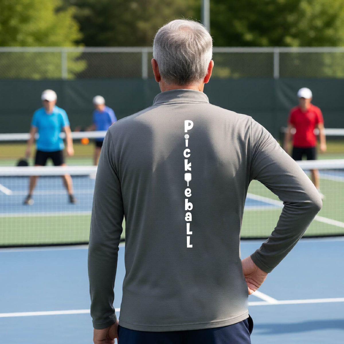 A man wearing a Gray long-sleeve quarter-zip shirt with a white graphic text 'PICKLEBALL' on the back with a pickleball paddle replacing the L, standing on a pickleball court.