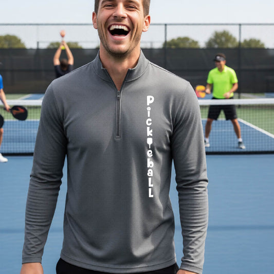 A man wearing a Gray long-sleeve quarter-zip shirt with a white graphic text 'PICKLEBALL' on the front with a pickleball paddle replacing the L, standing on a pickleball court.

