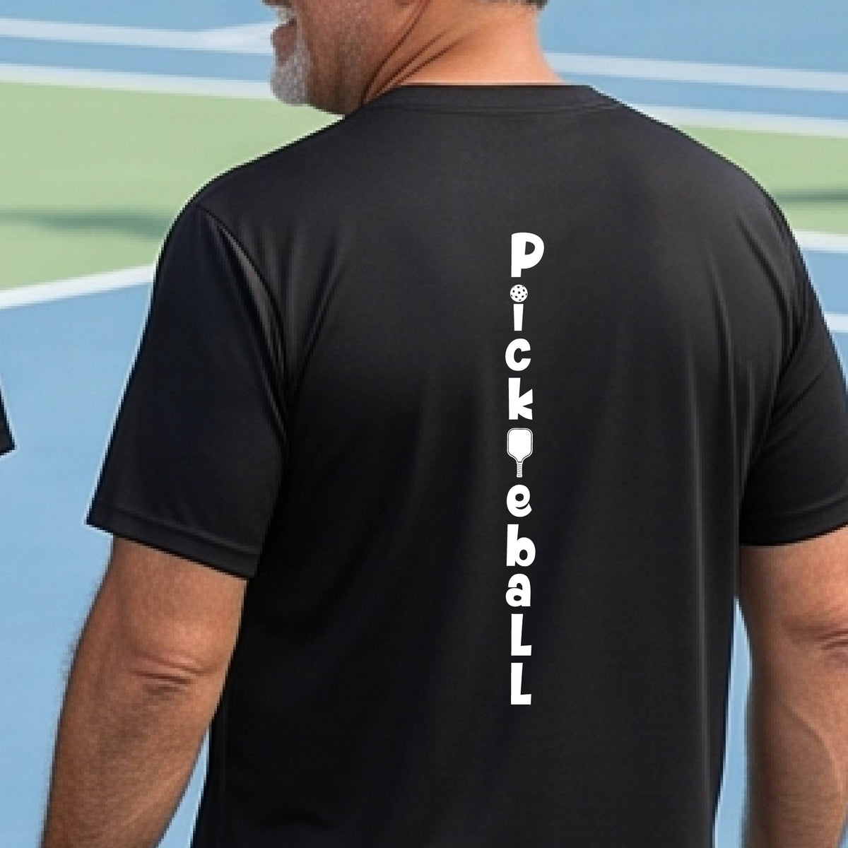 A man wearing a black short-sleeve t-shirt with a white graphic text 'PICKLEBALL' on the back with a pickleball paddle replacing the L, standing on a pickleball court.
